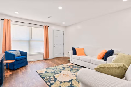 Bright living room with white sofa, colorful pillows, armchair, patterned rug, and peach curtains.