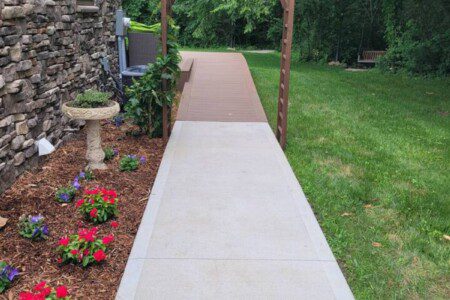 Concrete walkway leading to a wooden arbor beside a stone house, bordered by flowers and green lawn.