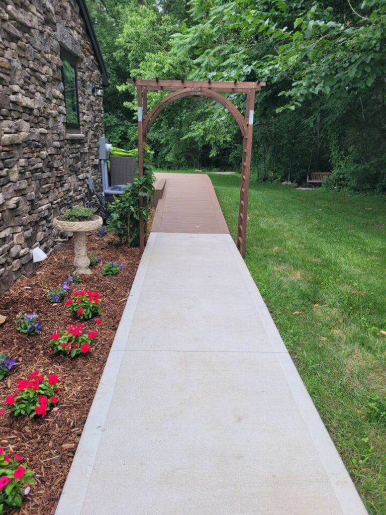 Concrete walkway leading to a wooden arbor beside a stone house, bordered by flowers and green lawn.