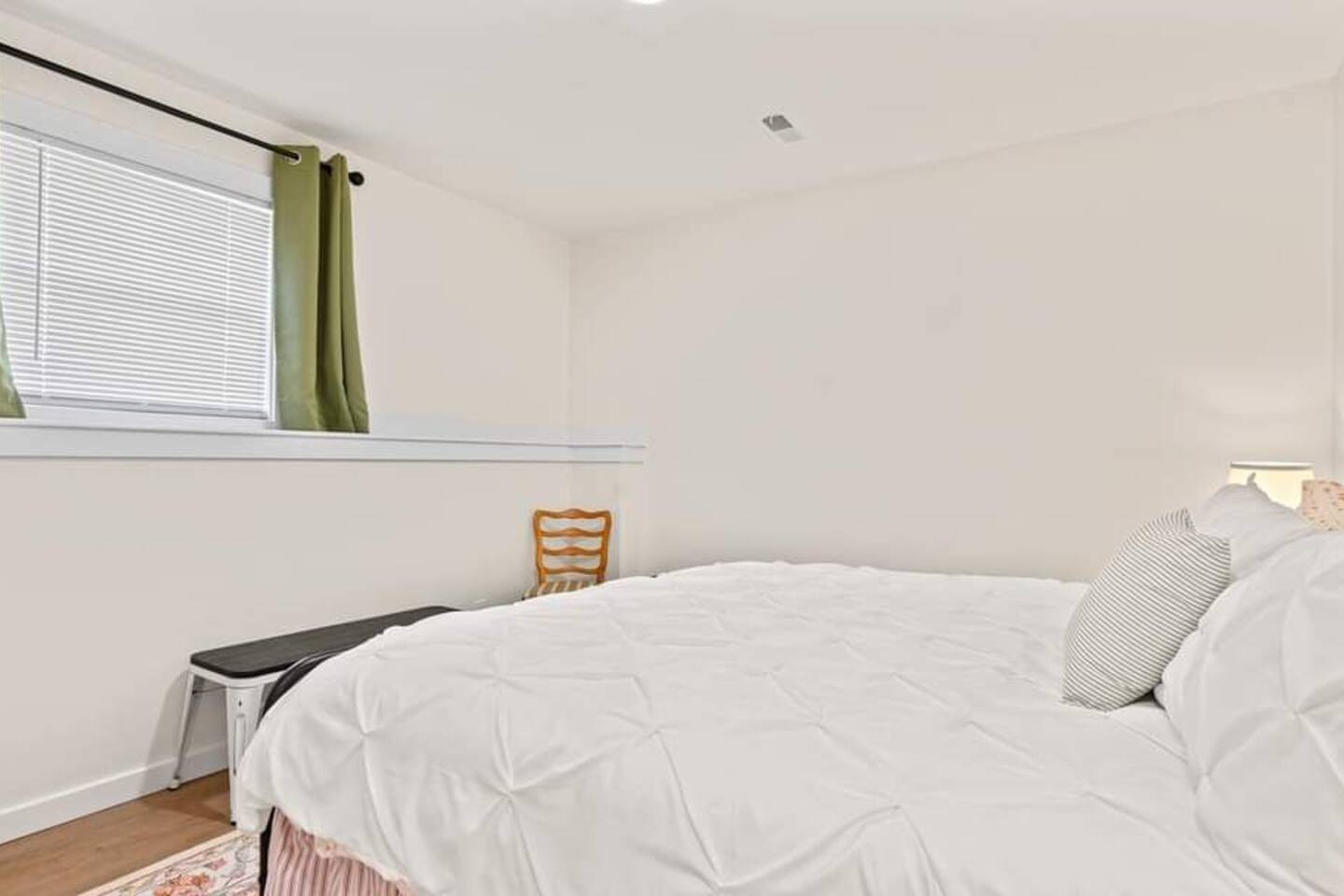Bright bedroom with a white comforter, striped accent pillows, wood flooring, a small bench at the foot of the bed, and a window with green curtains.