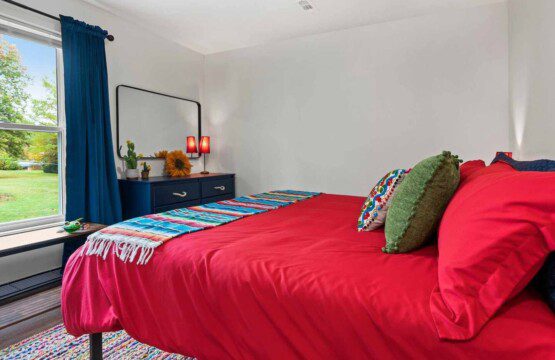 Bedroom with a red bedspread, colorful striped throw blanket, green accent pillows, blue curtains, and a dresser with a mirror and small lamps near a window overlooking a grassy yard.