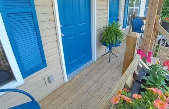 Charming front porch with bright blue doors and shutters, beige siding, black lantern-style wall lights, potted fern on a blue metal stand, and colorful flowers along the wooden railing.