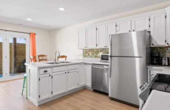 Bright kitchen with white cabinets, stainless steel refrigerator and dishwasher, double sink island, floral backsplash, and glass doors leading outside with coral curtains.