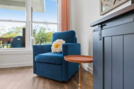 Blue upholstered armchair with a white floral accent pillow beside a small round side table, positioned near a bright window with outdoor views.