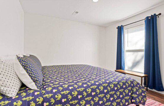 Bedroom with a queen bed dressed in a navy patterned comforter, white pillows, blue curtains, and a window with blinds letting in natural light.
