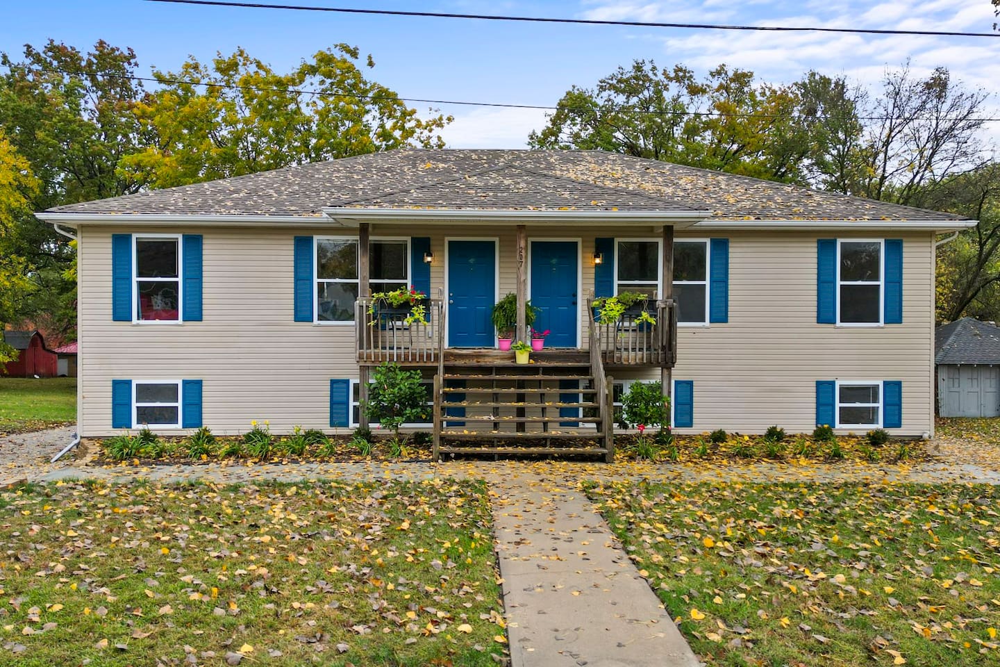 Two-story beige duplex with blue shutters, shared porch, potted plants, and autumn leaves on the lawn.