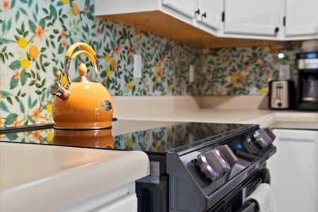 Close-up of a kitchen stove with stainless steel hood, white cabinets, colorful floral backsplash, and a yellow kettle on the cooktop.