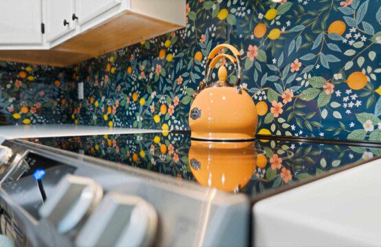 Yellow tea kettle on a glass-top stove with a floral citrus-patterned backsplash and white kitchen cabinets above.
