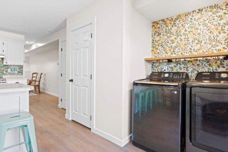 Modern laundry area with black washer and dryer against a floral wallpaper accent wall, light wood flooring, white doors, and a glimpse of the adjacent kitchen with mint green stools.