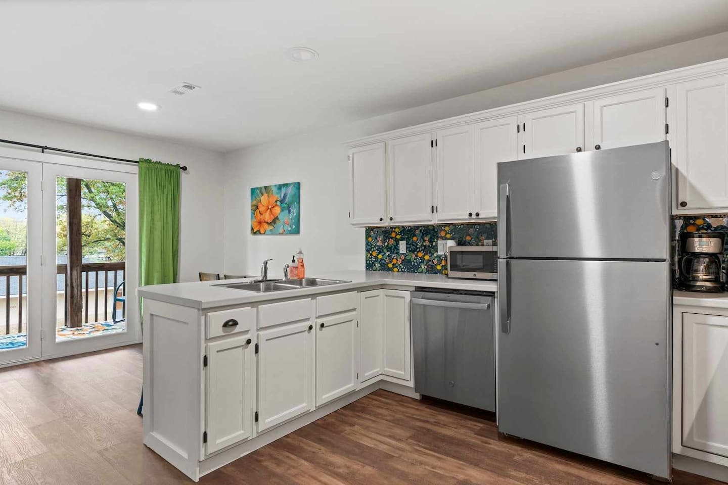 Bright white kitchen with stainless steel refrigerator and dishwasher, peninsula sink, citrus-patterned backsplash, wood floors, and glass doors leading to a balcony with green curtains.