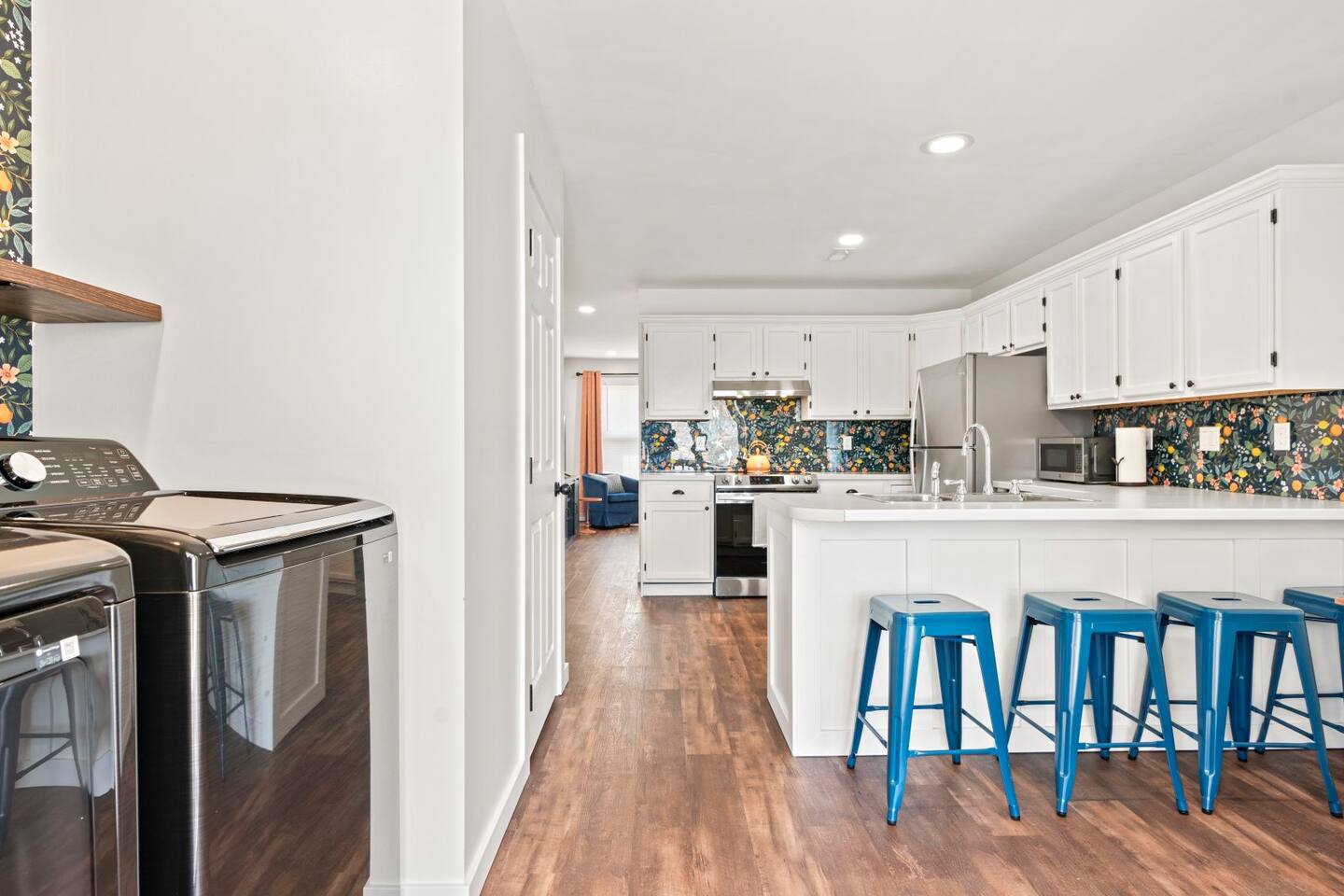 Bright, modern kitchen with white cabinets, a floral-patterned backsplash, stainless steel appliances, a center island with blue metal stools, and wood-style flooring.