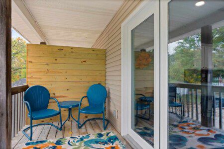 Small covered balcony with two blue chairs and a matching side table, wood privacy wall, sliding glass door, and patterned outdoor rug.