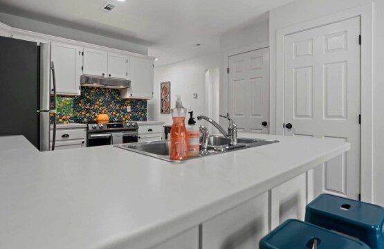 Modern white kitchen with peninsula sink, stainless steel appliances, colorful citrus-patterned backsplash, and teal bar stools.