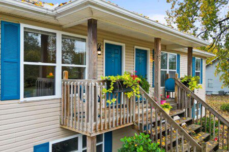 Duplex entrance with two front doors, shared porch, potted plants, blue chair, and autumn trees in the background.