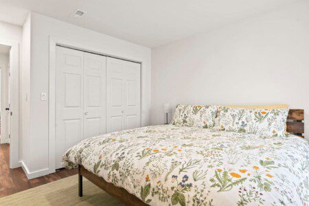 Spacious bedroom with a wooden platform bed featuring white botanical bedding, sliding closet doors, and a small bedside lamp.