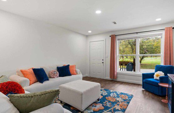 Park place 1 for extended stay: Bright living room with a cream sectional sofa, coral and navy accent pillows, matching ottoman, blue armchair, floral area rug, and large window with peach curtains overlooking a green yard.