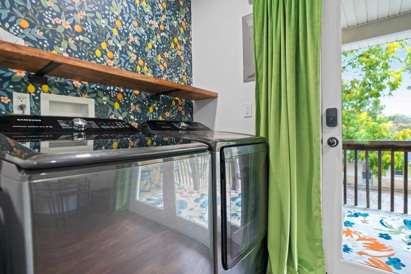 Laundry area with a front-loading washer and dryer, wooden shelf above, floral wallpaper, and a glass door with green curtains leading to a balcony.