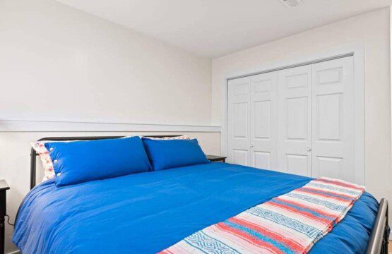 Bedroom with a blue bedspread and matching pillows, striped throw blanket at the foot of the bed, bedside tables, and double closet doors.