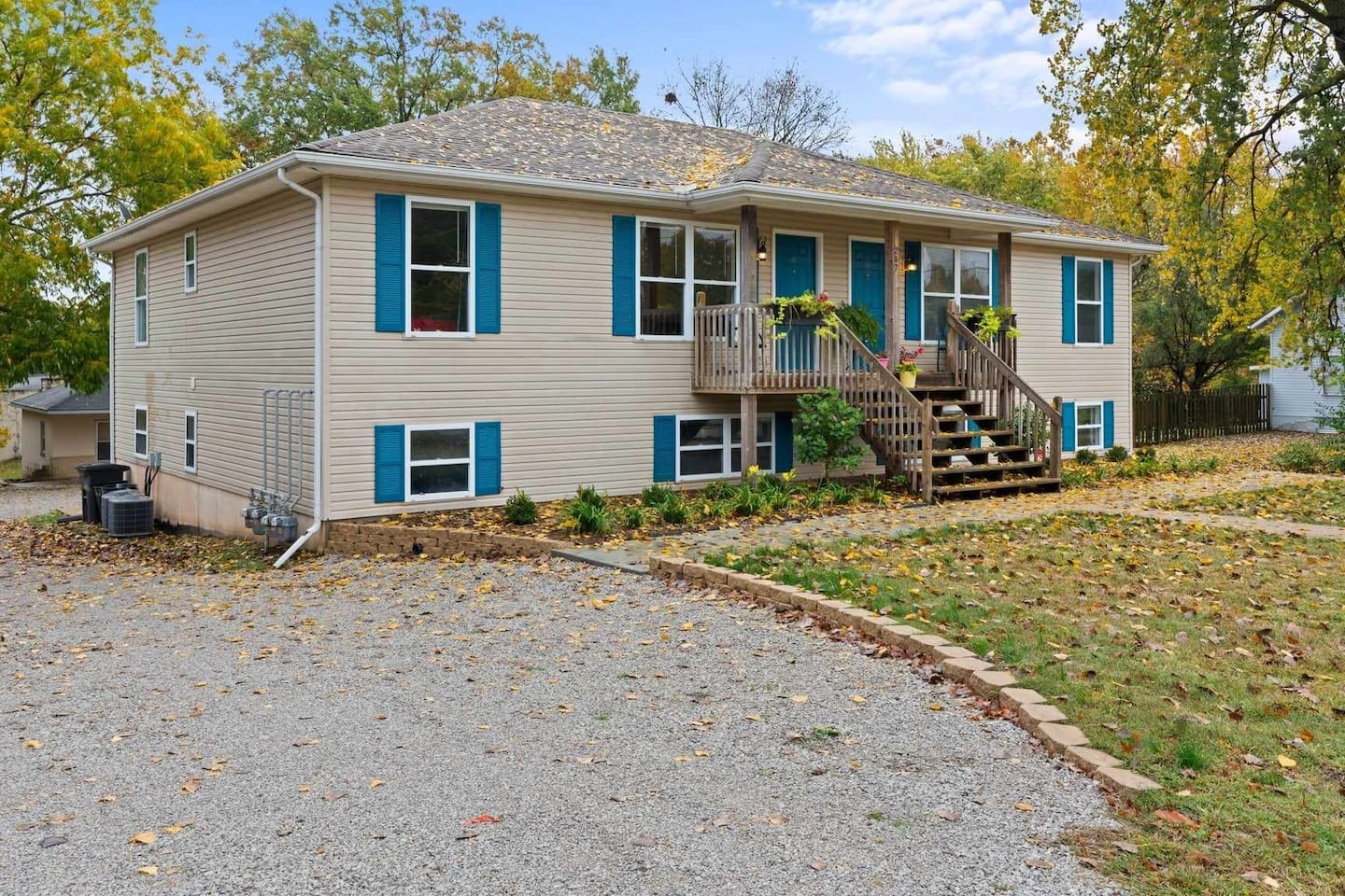 Exterior view of a two-level apartment building with blue doors, wooden steps, and a gravel driveway surrounded by trees