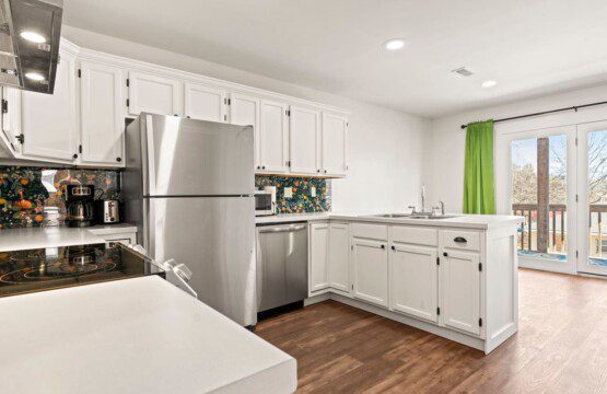 Bright kitchen with white cabinets, stainless steel refrigerator and dishwasher, floral-patterned backsplash, L-shaped counter with sink, wood flooring, and glass doors with green curtains leading outside.