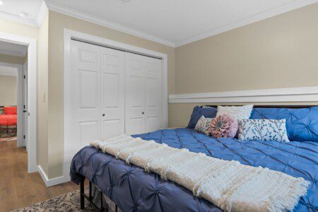 Bedroom with a blue quilted bedspread, decorative accent pillows, white closet doors, and an open doorway leading to a second bedroom with coral bedding.