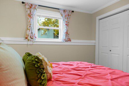 Bedroom with a coral quilted bedspread, green and white accent pillows, bedside lamp, floral curtains, and white sliding closet doors.