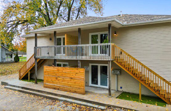 Modern raised home exterior with upper-level balcony, natural wood staircases on both sides, privacy fence panel, and concrete patio surrounded by autumn trees.
