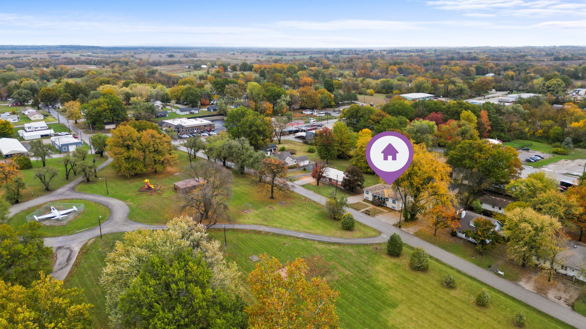 Aerial neighborhood view in autumn with colorful trees, curved streets, a small park with playground equipment and an airplane display, and a highlighted home marked with a location pin icon.