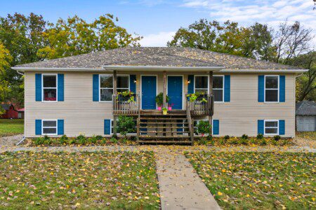 Front-facing symmetrical raised duplex with beige siding, bright blue doors and shutters, central wooden staircase, porch planters, and a leaf-covered lawn in autumn.